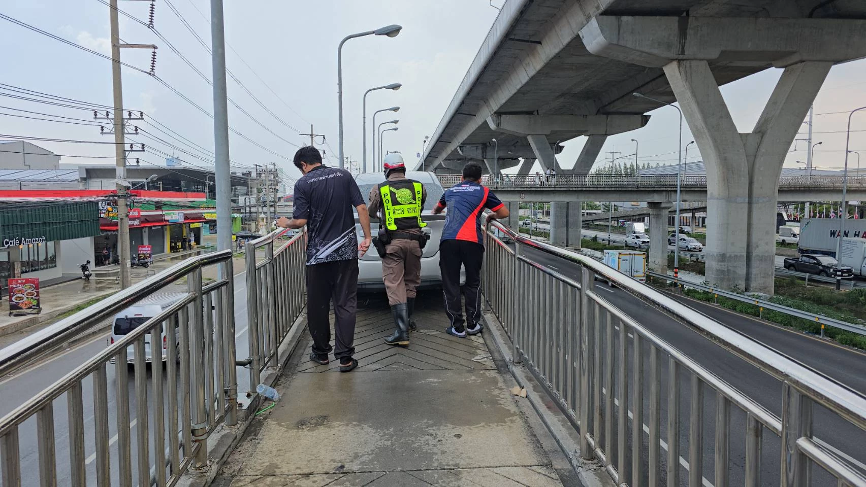 Drei Personen, darunter ein Polizist in gelber Warnweste, stehen hinter einem weißen Fahrzeug auf der überdachten Brücke in Samut Sakhon