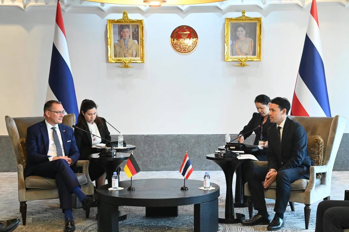Zwei Gesprächspartner sitzen in Sesseln vor thailändischen Flaggen und Porträts, auf dem Tisch stehen eine deutsche und eine thailändische Tischflagge